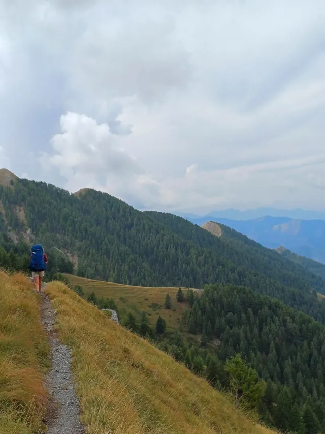 Un randonneur sur un sentier de crête, entre les pentes boisées et les nuages.